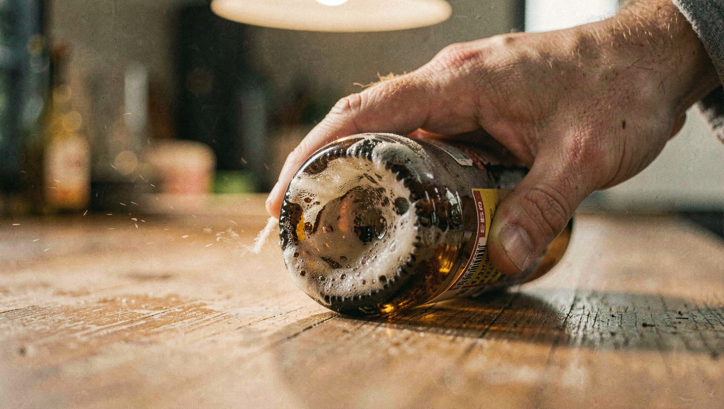 A hand rolling a brown beer bottle on its side across a wooden counter, cloudy sediment swirling in the last inch of beer, warm overhead light