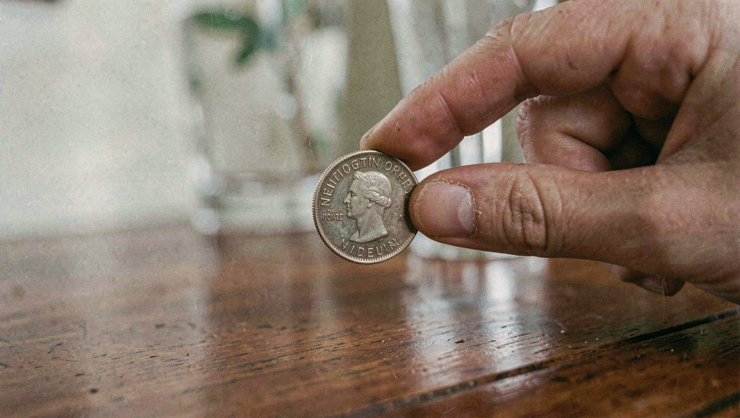 A close crop of a hand holding a milled edge coin flat between thumb and forefinger a few centimetres above a lacquered table, glass out of focus behind