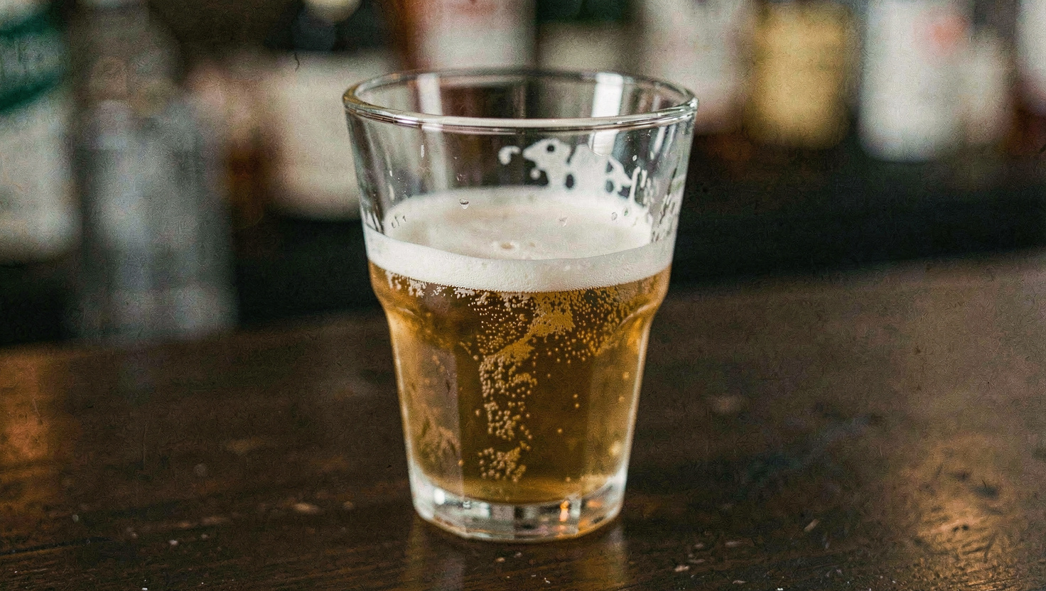 Close crop of the upper third of a nonic pint glass on a dark bar top showing the bulge below the rim, lacing on the inside wall, warm light from one side