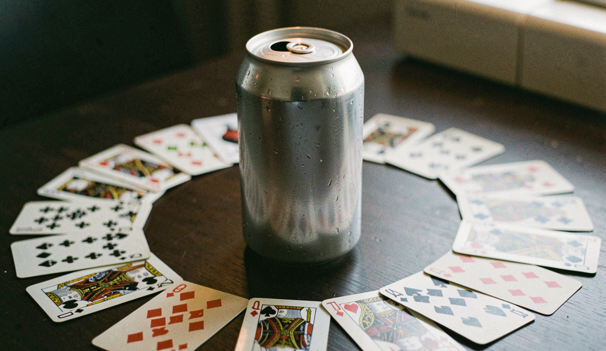 An unopened can standing upright in the middle of a ring of face down playing cards on a dark table, condensation on the metal, lamp reflected on the lid